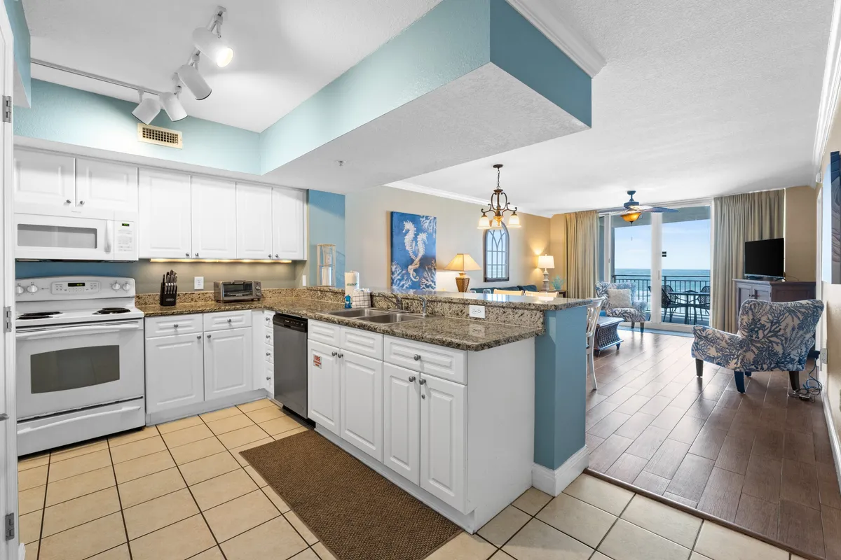 A kitchen scene featuring granite countertops and white cabinetry, with large windows offering ocean views in the background. The atmosphere is bright and cheerful, blending the kitchen’s functional appeal with a sense of vacation relaxation.