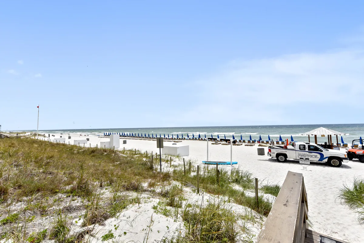 A wide stretch of sandy beach is dotted with colorful umbrellas and lounge chairs, all facing the sparkling ocean. The golden light hints at a sunset, inviting guests to unwind and soak in the view.