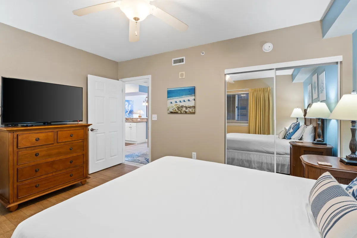 A calm and cozy second bedroom featuring a queen-size bed, wooden dresser, television, mirrored wardrobe, and coastal artwork. Sunlight filters in, creating a welcoming and restful ambiance.