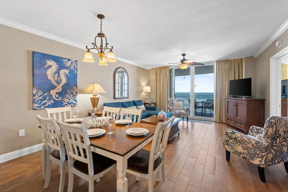 A cozy dining area is shown with a dining table set for a meal, seahorse artwork on the wall, a blue sofa nearby, and a balcony visible through large windows. The space is bright, inviting, and highlights the connection between dining, living, and the outdoor balcony area, reinforcing the theme of togetherness with sunset views.