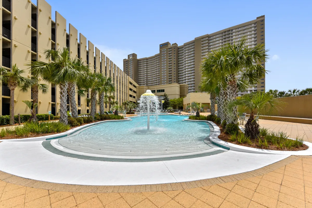 A vibrant exterior view highlighting a sparkling resort-style swimming pool, surrounded by palm trees and lounge chairs, with the building facade visible in the background. The setup invites socializing, relaxing, and play for all ages.