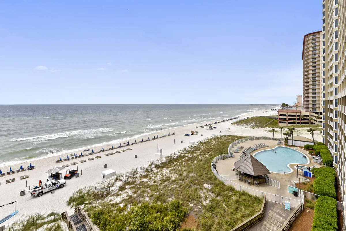 This image captures the resort's beachfront location, showing both the pristine beach and the adjacent swimming pool area. Lounge chairs are neatly arranged for relaxation, and the tranquil ocean provides a scenic backdrop, while the buildings hint at the convenience of being steps away from family-friendly amenities.